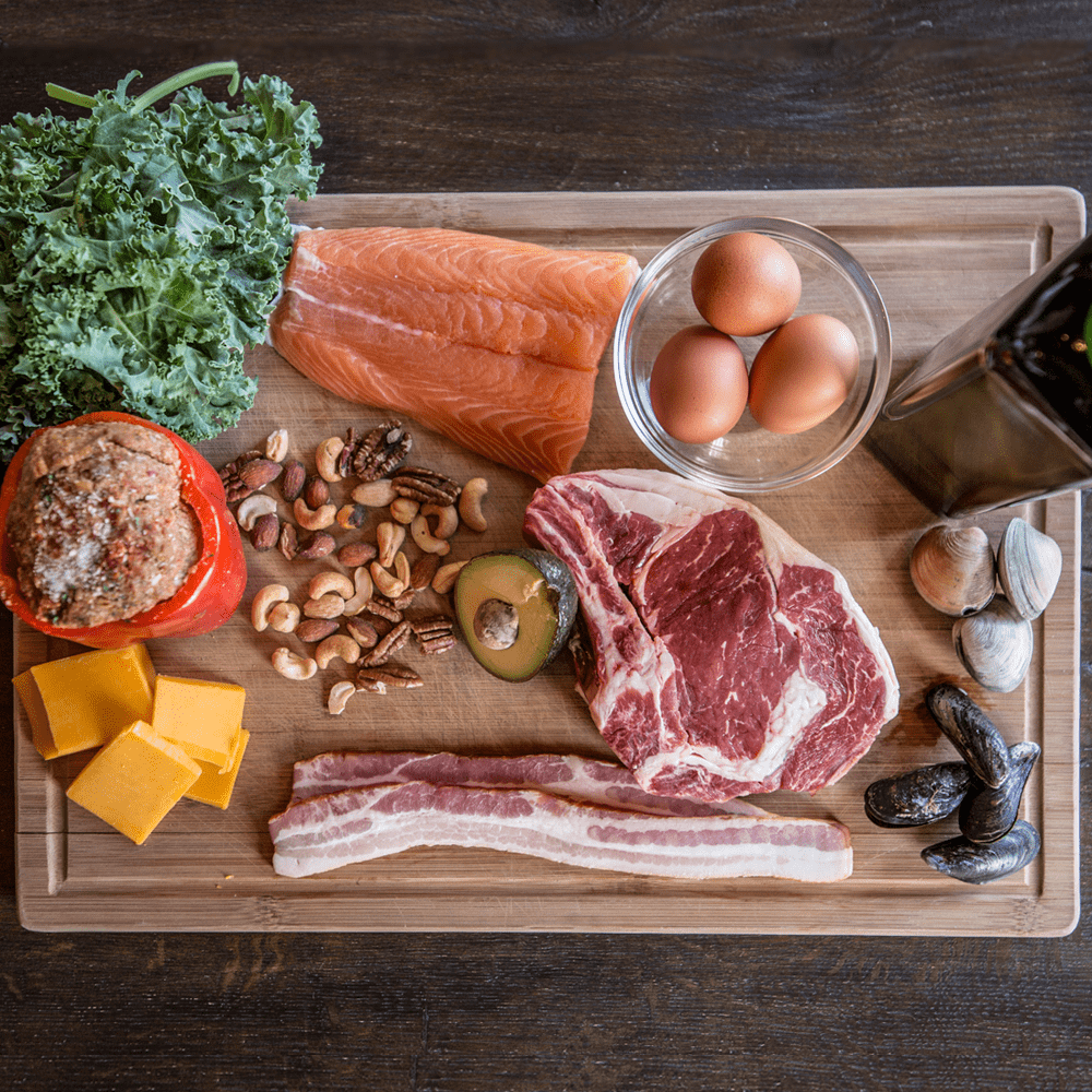 Whole foods on a cutting board: salmon, eggs, steak, kale, nuts, avocado, cheese and shellfish