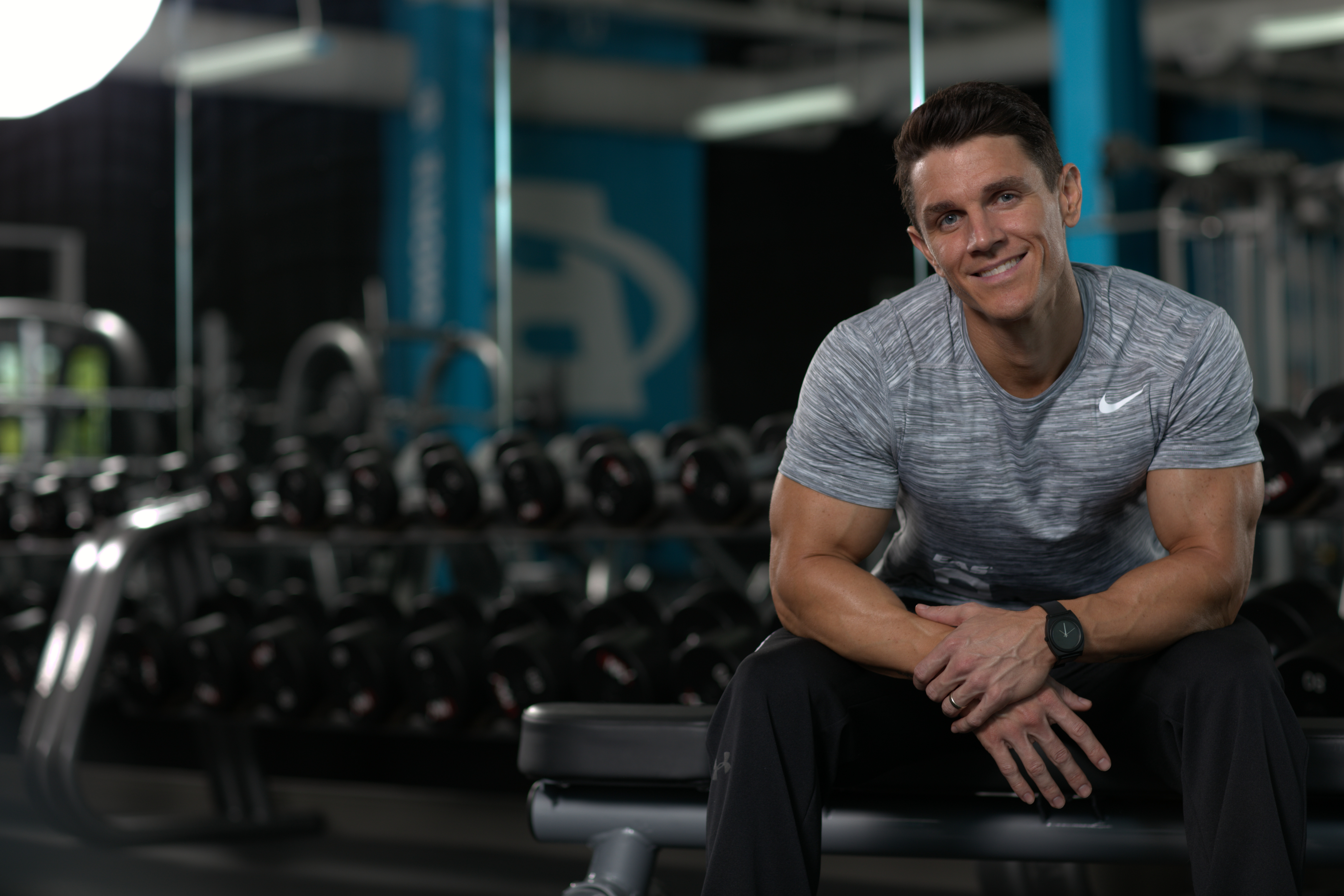 Jason Wittrock smiling, seated on a weight bench in front of a rack of dumbbells